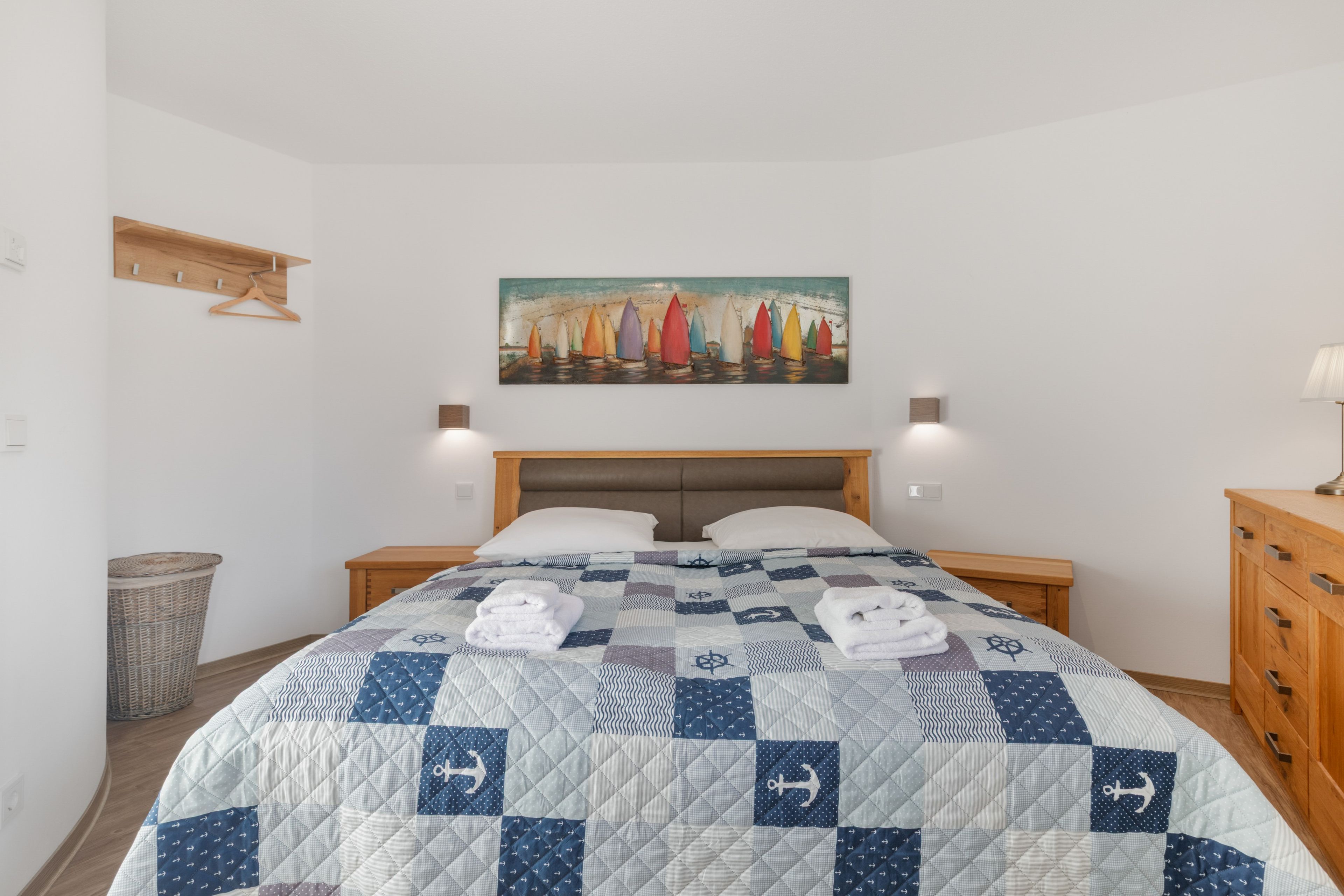 A hotel room with a bed, a painting of sailboats, a wooden dresser, and a nightstand.
