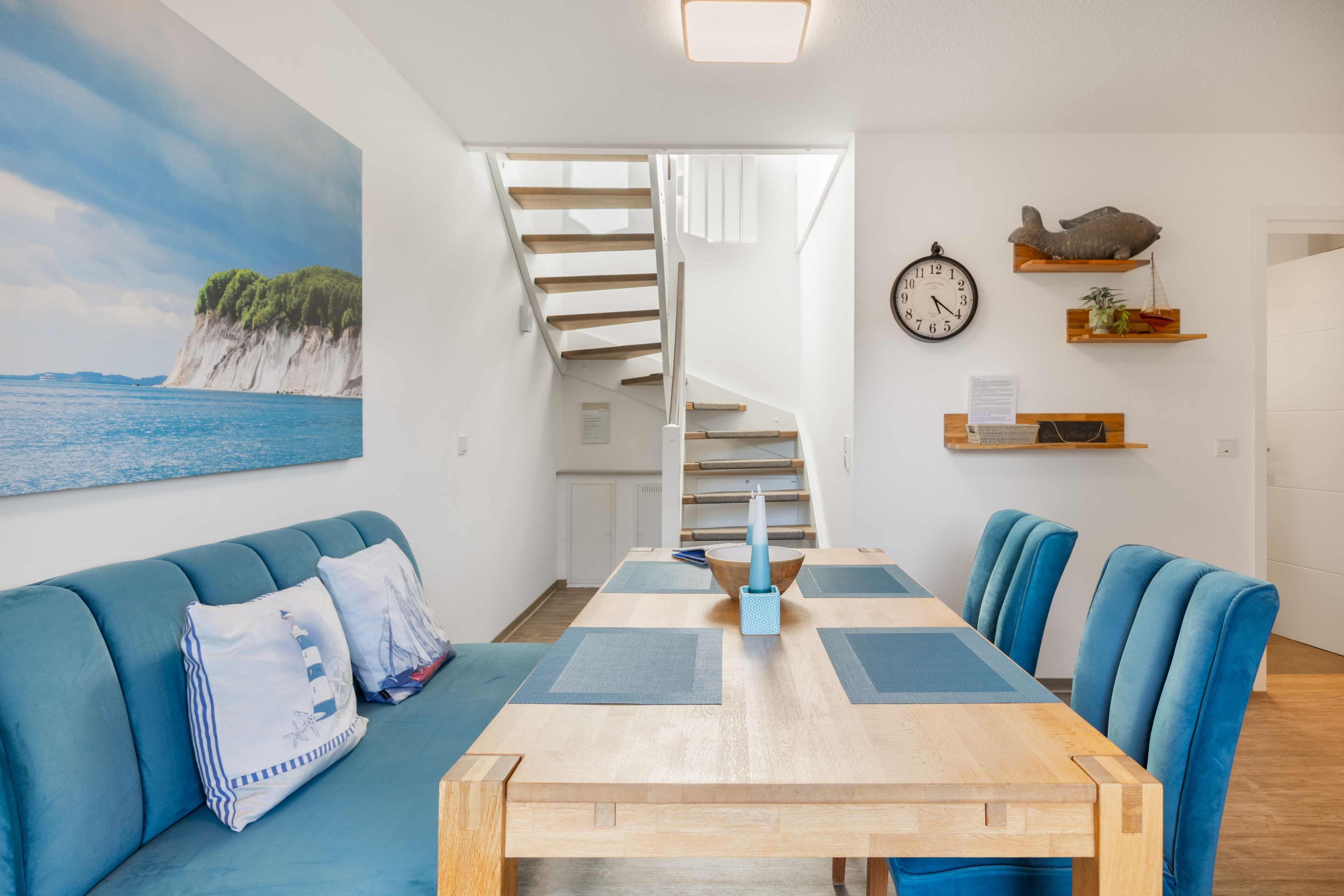 A modern living room with a wooden dining table, blue upholstered chairs, a blue sofa with patterned cushions, a spiral staircase, and a view of the sea through a large window.