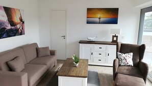 A modern living room with a sofa, armchair, coffee table, and a painting of a sunset over water. - Enjoy Sea Views & Tranquility – Exclusive Penthouse Apartment in Lauterbach Harbor (Putbus)