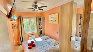 Een slaapkamer met een bed, een plafondventilator, een raam met gordijnen, een plank met manden, en een schilderij aan de muur.