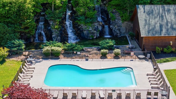 A swimming pool with lounge chairs, a wooden cabin, and a waterfall. - Pet-Friendly | Hot Tub | Outdoor TV | Game Room | Views | Sleeps 8 (Sevierville)