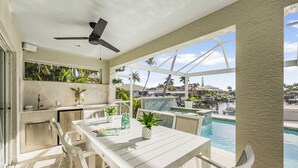 Un área de comedor exterior moderna con vista a la piscina, mobiliario blanco y un ventilador de techo.