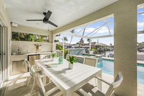 Ein moderner Essbereich im Freien mit Blick auf einen Pool, weißen Möbeln und einem Deckenventilator.