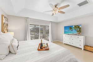 Ein Schlafzimmer mit einem Bett, einem Deckenventilator, einem Fernseher, einer Kommode und einem Fenster mit Vorhängen.