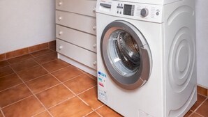 Ein kleiner Waschraum mit einer Waschmaschine mit Frontlader, einer hohen Kommode mit Schubladen und einem oberhalb angebrachten Regal für Reinigungsmittel.