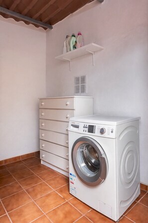 A compact laundry room with a front-loading washing machine, a tall dresser with multiple drawers, and a shelf above holding cleaning supplies. - TenoHomes Cozy Rustic Home with Terrace & Views (Buenavista del Norte)