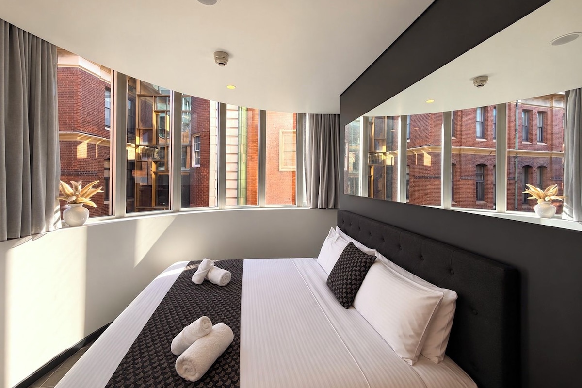 A modern hotel room with a large bed, a view of a brick building, and a security camera on the ceiling.