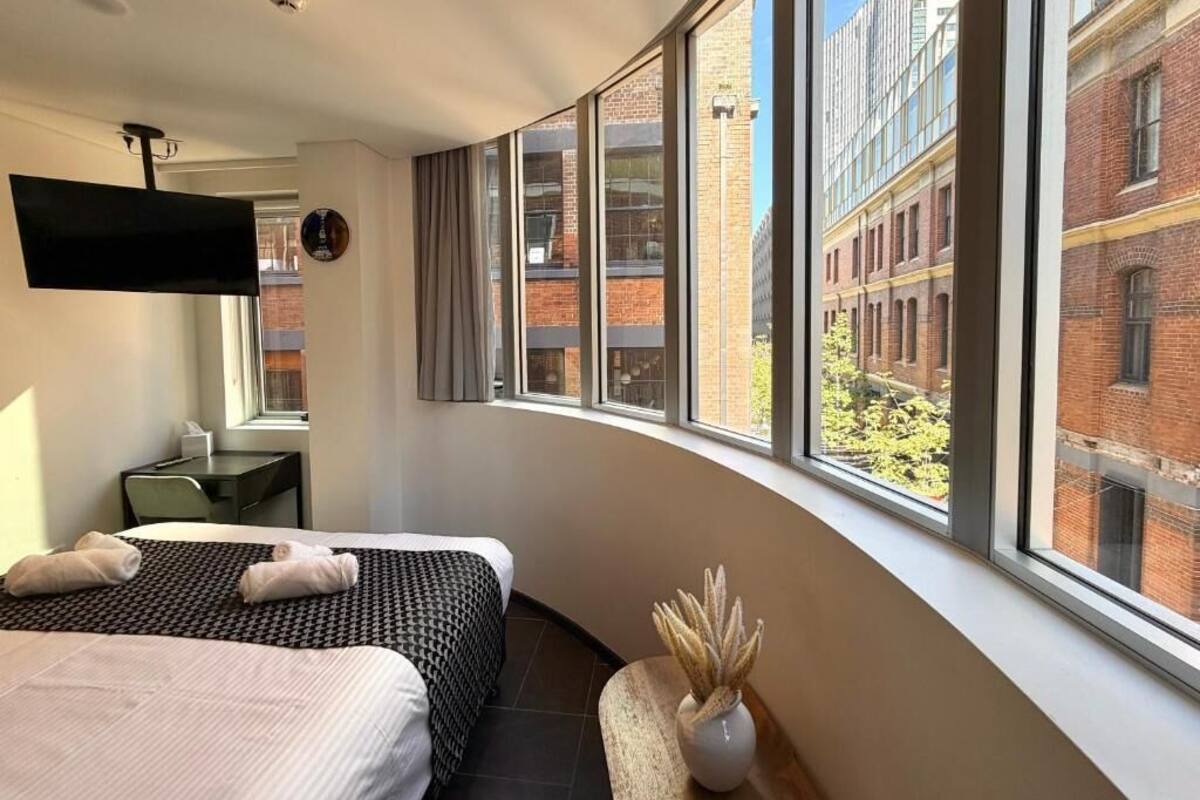 A hotel room with two beds, a desk, a flat-screen TV, and large windows with a view of brick buildings.