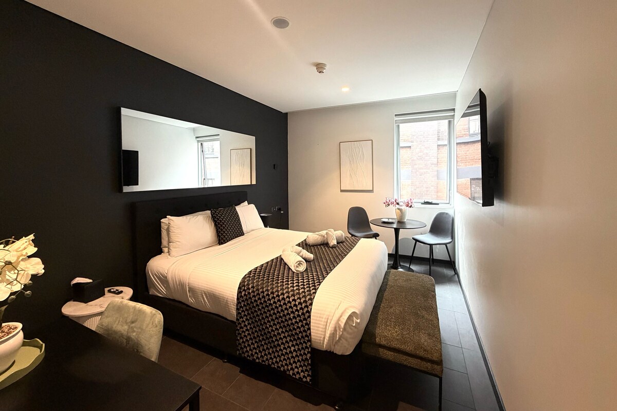 A modern hotel room with a large bed, a desk, a chair, a small table, and a window.