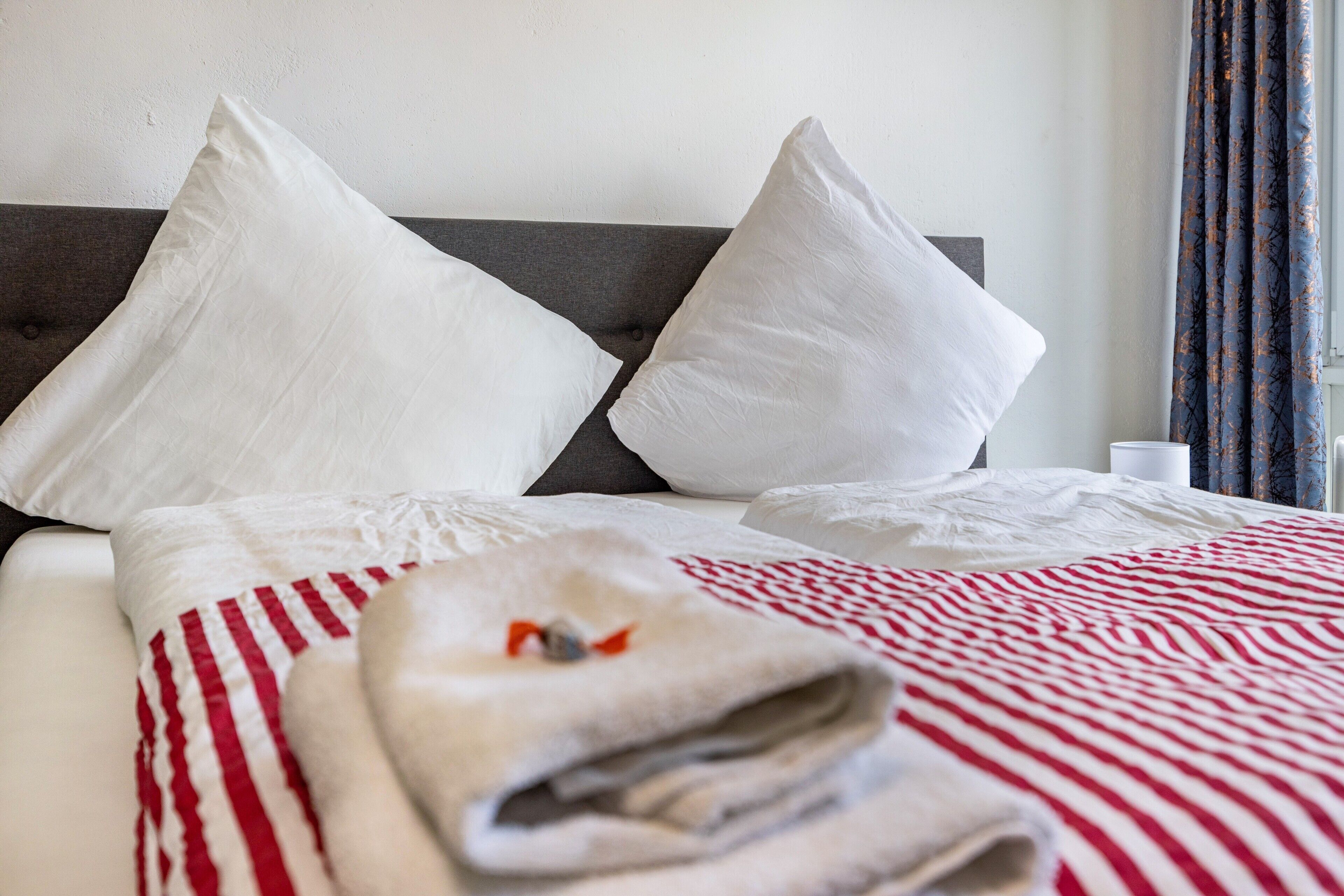 Een net opgemaakt bed met witte kussens, een gestreepte rode en witte dekbedovertrek, en een opgevouwen handdoek op het bed.