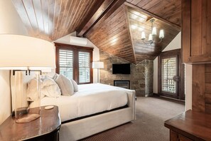 Ein Schlafzimmer mit Bett, Fernseher, einer Steinwand, Holzbalken an der Decke und einem Fenster mit Jalousien.
