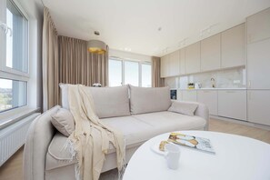 A modern living room with a beige sofa, a round white table, and a kitchen area with white cabinets. - Elegant Apartment | Parking, Balcony, Air Conditioning (Warszawa)