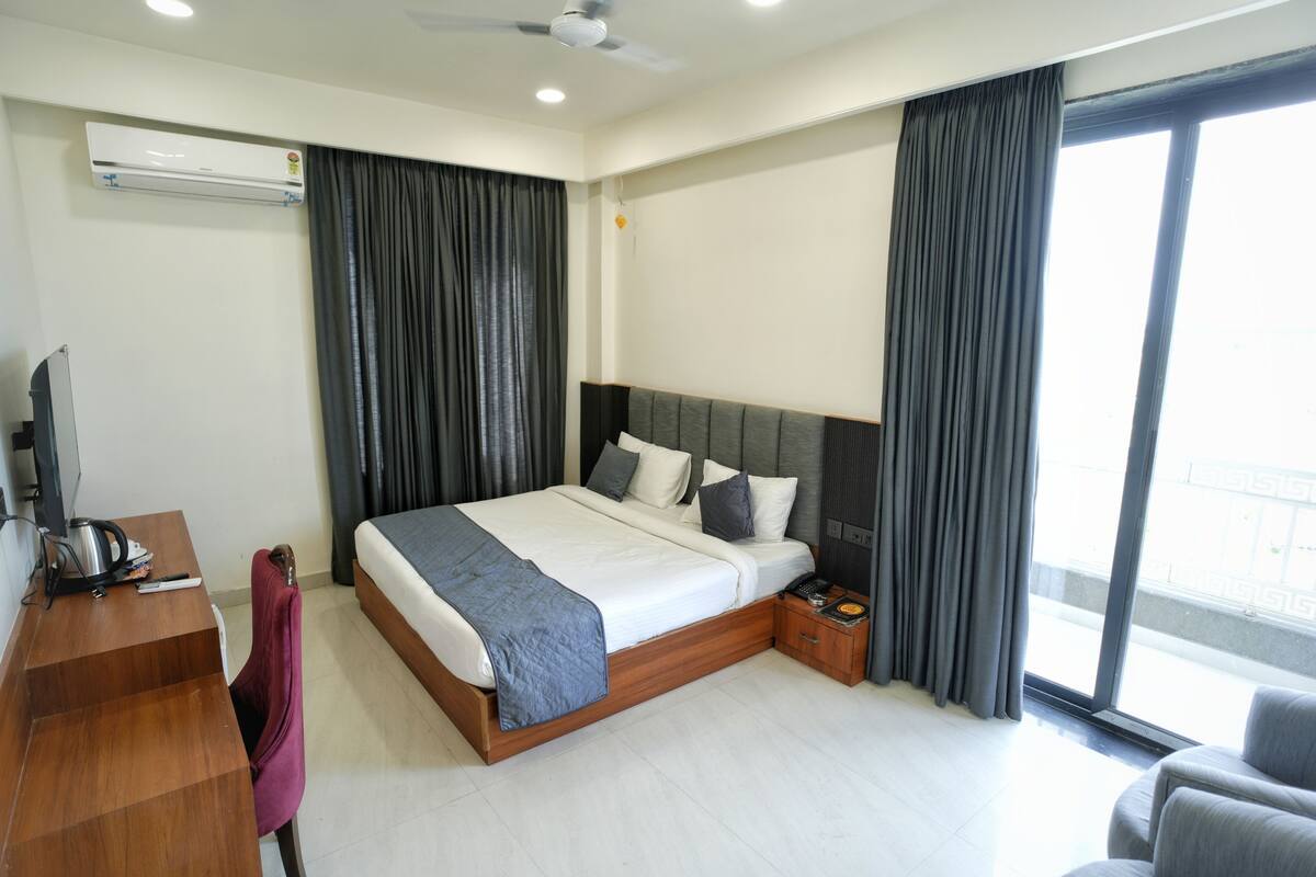 Habitación de hotel con cama, escritorio, silla, televisor, aire acondicionado y ventana con cortinas.