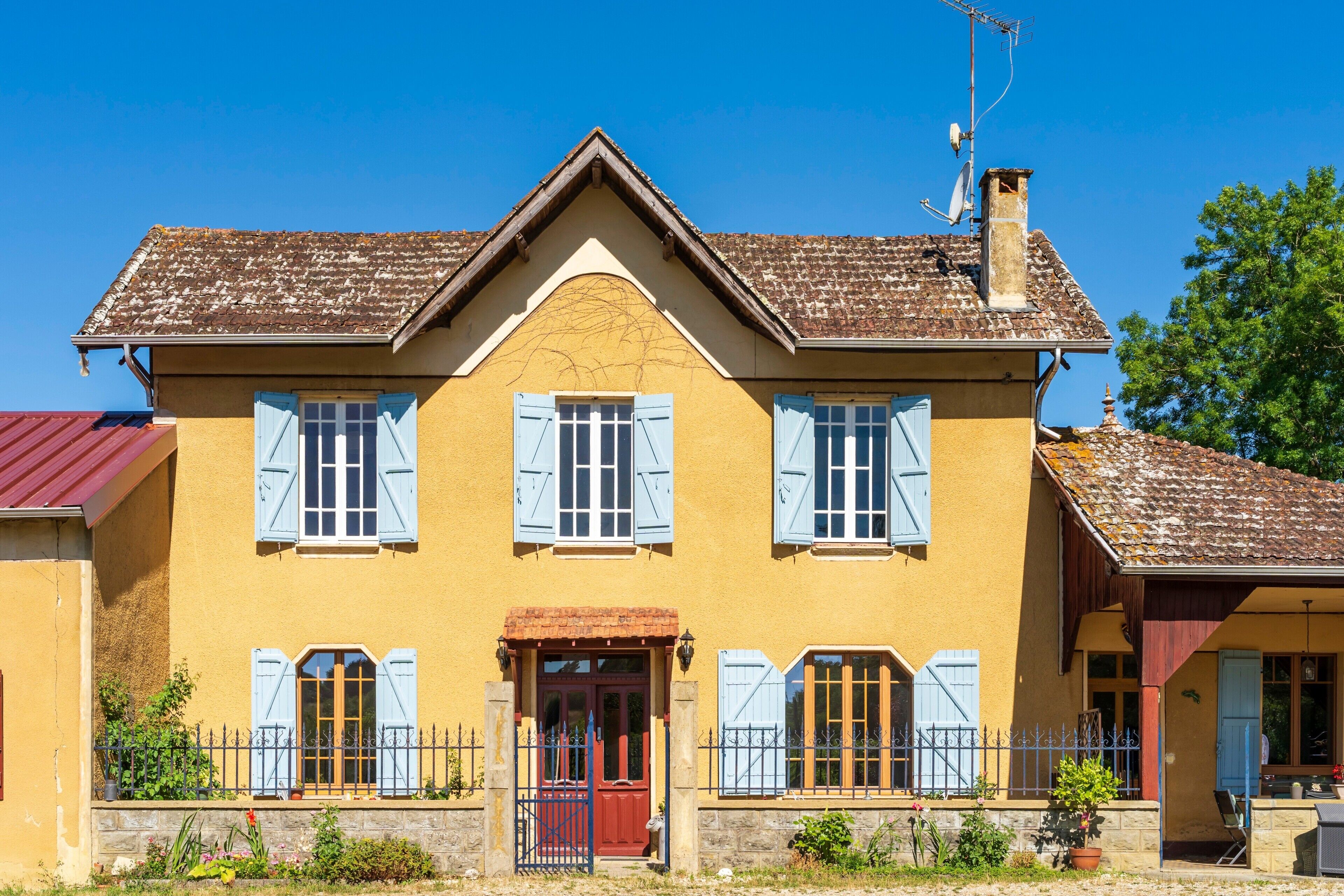 Une maison à deux étages avec une porte rouge, des volets bleus et un toit recouvert de tuiles.