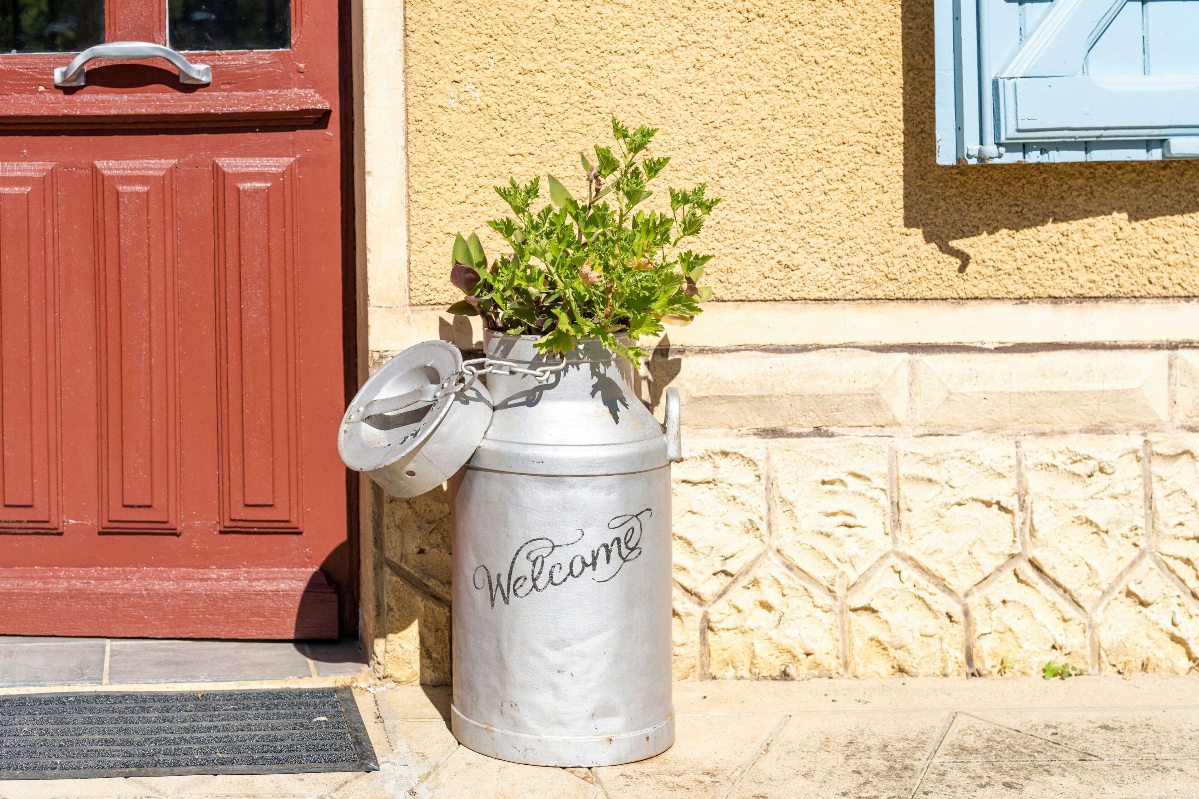 Un récipient en métal portant l’inscription « Bienvenue », placé sur une surface en pierre à côté d’une porte rouge.