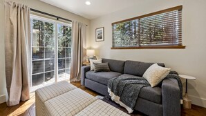 A living room with a grey sofa, a patterned ottoman, a side table with a lamp, a window with blinds, and a sliding glass door leading to an outdoor area. - The “Pine Tree Cabin”. Hot Tub, 4Br, 2 Ba, A/C, Near beaches, casinos, skiing (Stateline)