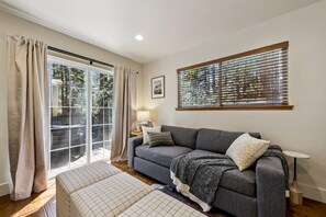 A living room with a grey sofa, a patterned ottoman, a side table with a lamp, a window with blinds, and a sliding glass door leading to an outdoor area. - The “Pine Tree Cabin”. Hot Tub, 4Br, 2 Ba, A/C, Near beaches, casinos, skiing (Stateline)