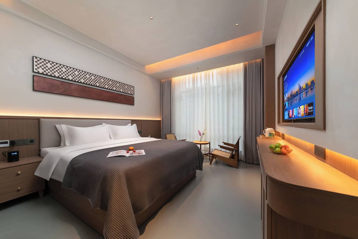 A modern hotel room with a large bed, bedside table, TV, and a small seating area.