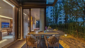 Un espacio moderno para comer al aire libre con mesa y sillas de madera, un ventanal de vidrio que da a una habitación y vista a un edificio residencial de fondo.