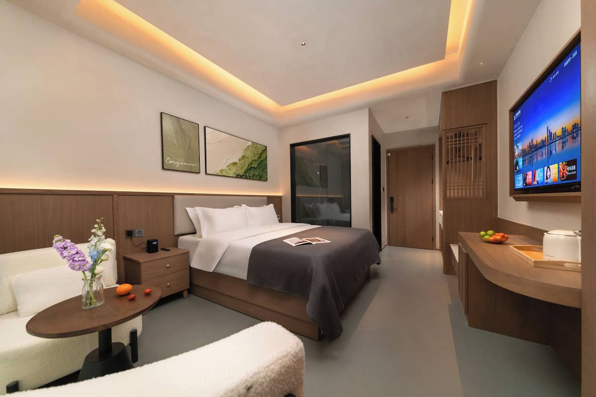 A modern hotel room with a bed, bedside table, TV, and a small seating area.