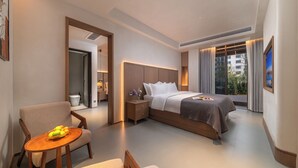 Habitación de hotel con cama, una silla, una mesita pequeña con fruta, una mesita de noche y un ventanal con cortinas.