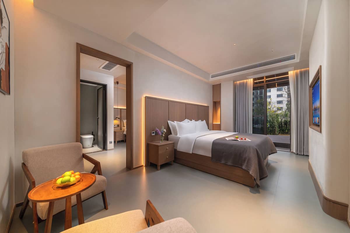 A hotel room with a bed, a chair, a small table with fruit, a nightstand, and a window with curtains.