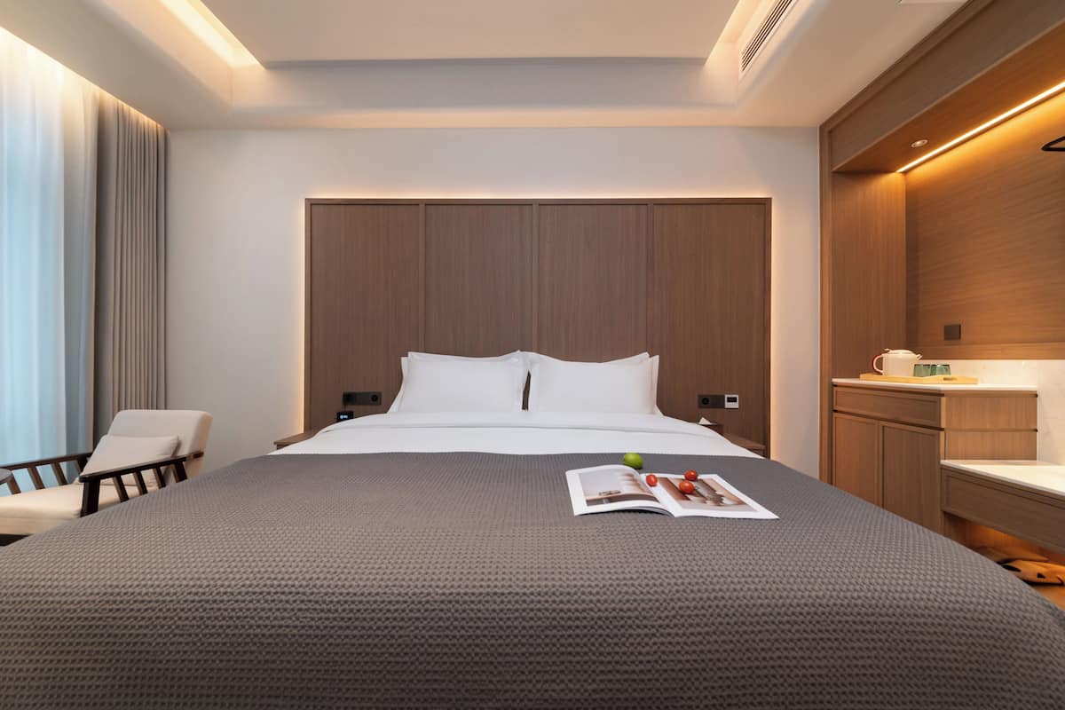 A modern hotel room with a large bed, a bedside table, a chair, and a built-in desk with a mirror.
