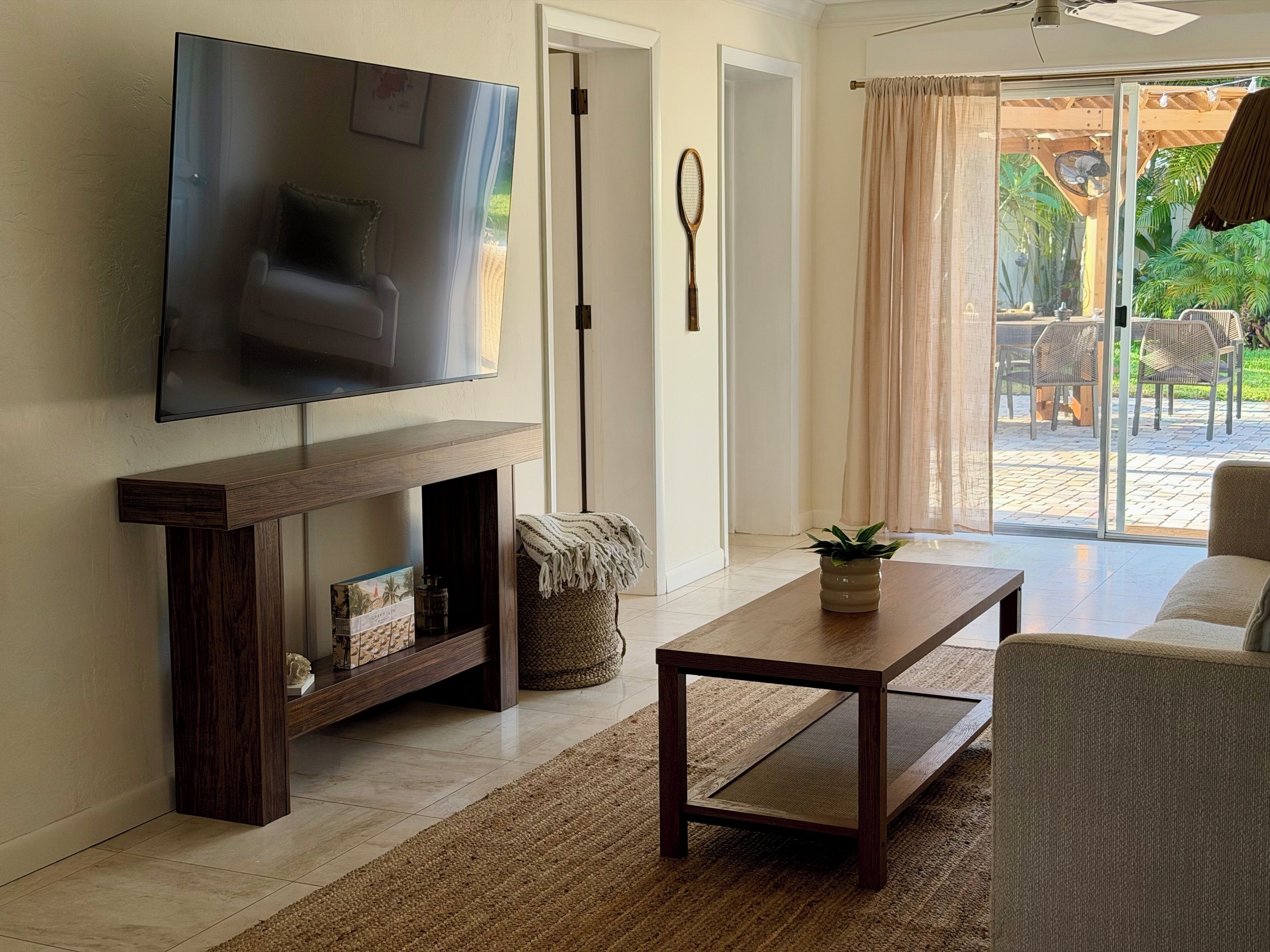 A living room with a flat-screen TV, a dark wooden coffee table, a woven pouf, and a potted plant.