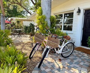 Ein Fahrrad mit Körben steht vor einem Haus mit einer gepflasterten Terrasse und einem Tisch mit Stühlen.