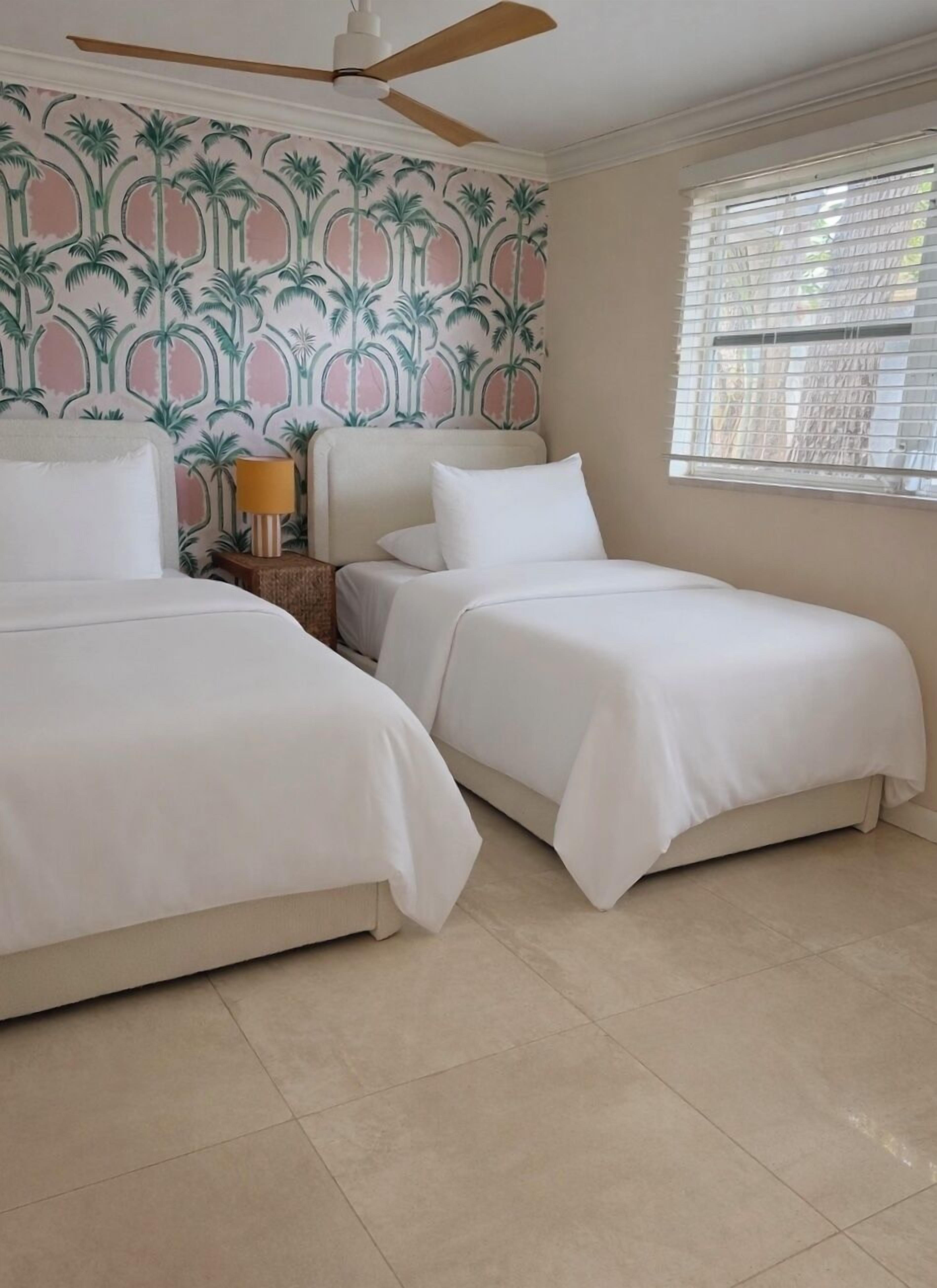 Habitación de hotel con dos camas, ventilador de techo, ventana con persianas y papel tapiz con diseño de palmeras.
