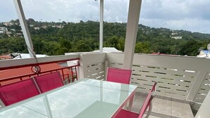 Une table à plateau en verre, entourée de chaises rembourrées de couleur rose, sur une terrasse carrelée offrant une vue sur une colline verdoyante.