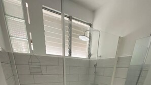 Une salle de bain avec une douche à parois de verre, des murs recouverts de carreaux blancs, un tapis rouge et un meuble-lavabo blanc avec un miroir.