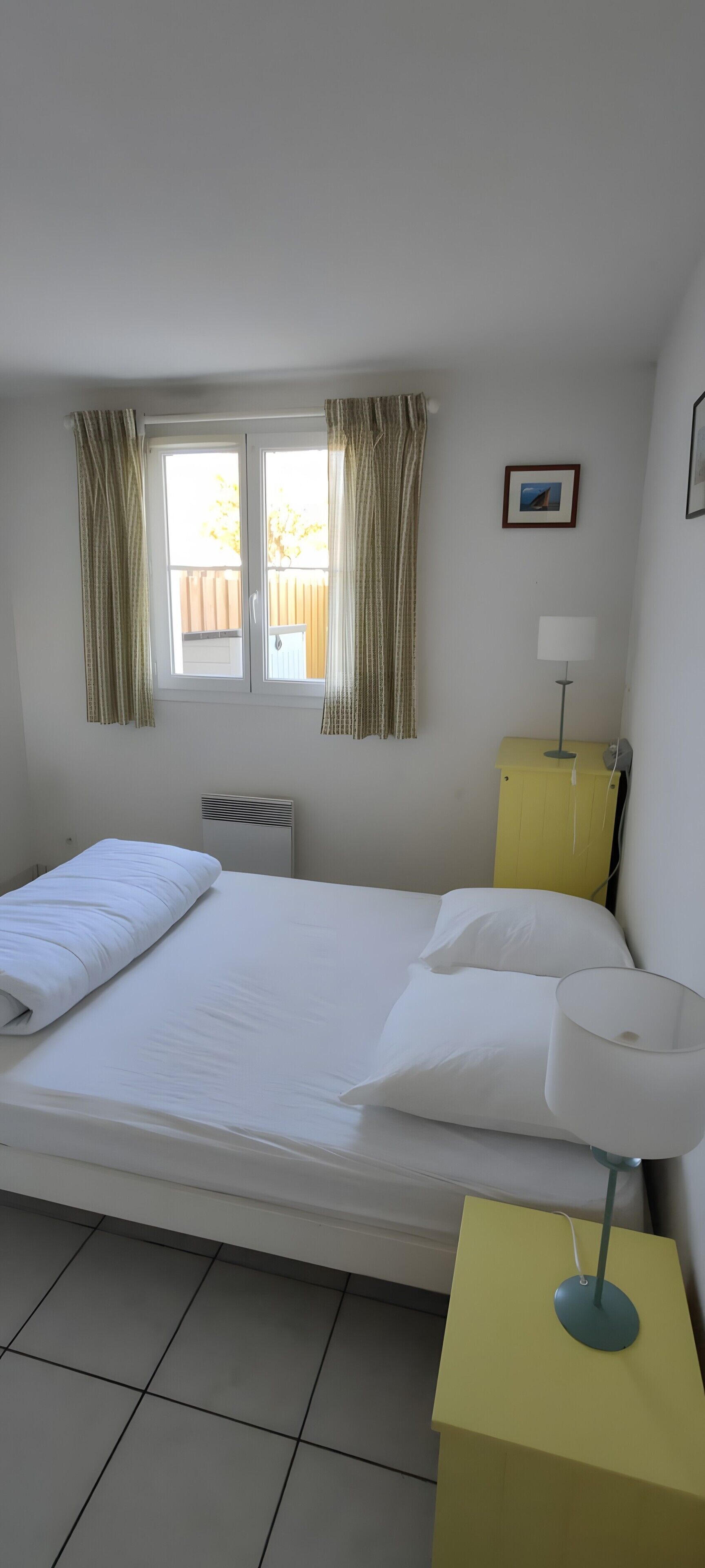 Ein ordentlich bezogenes Bett mit weißen Leinentüchern, ein Nachttisch mit Lampe, ein Fenster mit Vorhängen und ein kleines Bild an der Wand.