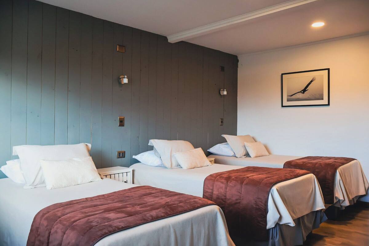 Quarto com três camas, cada uma com cabeceira branca e cobertor castanho. Há uma moldura com uma imagem na parede.