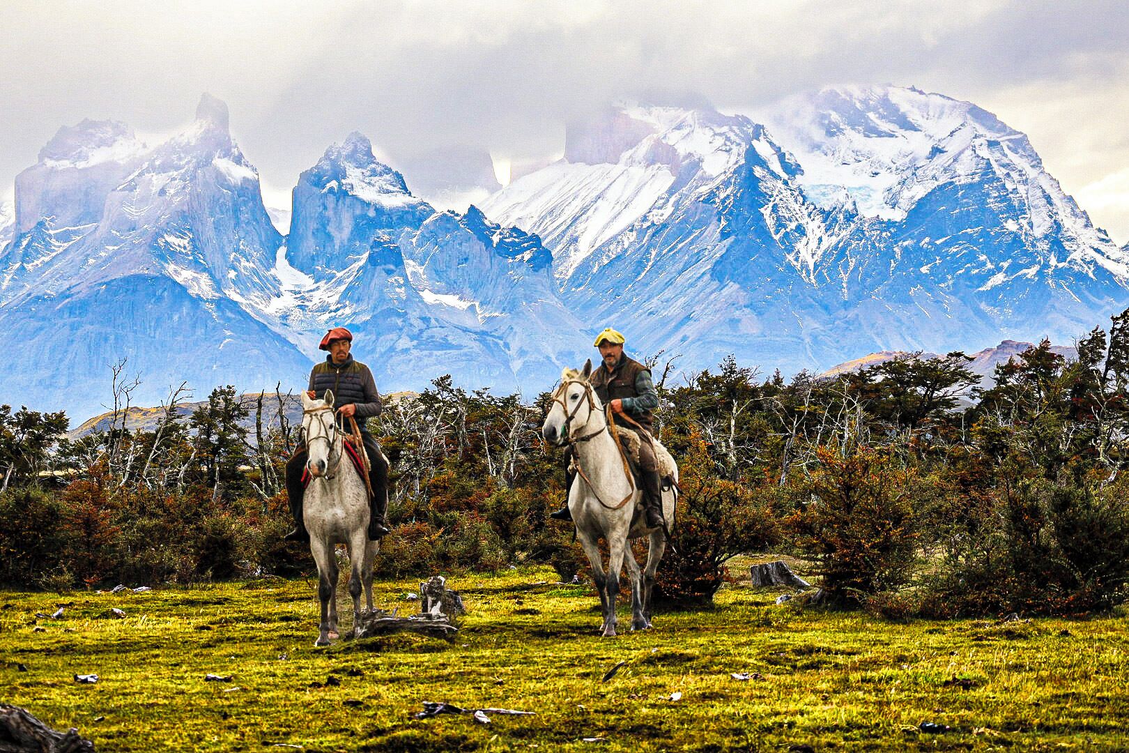 Two individuals on horseback in a grassy field with snow-capped mountains in the background.