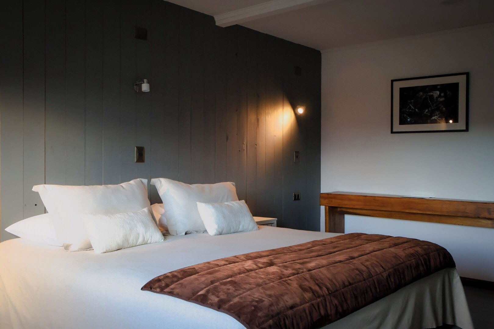 Quarto com cama, travesseiros, mesa de cabeceira, luminária na parede e cabeceira de madeira.