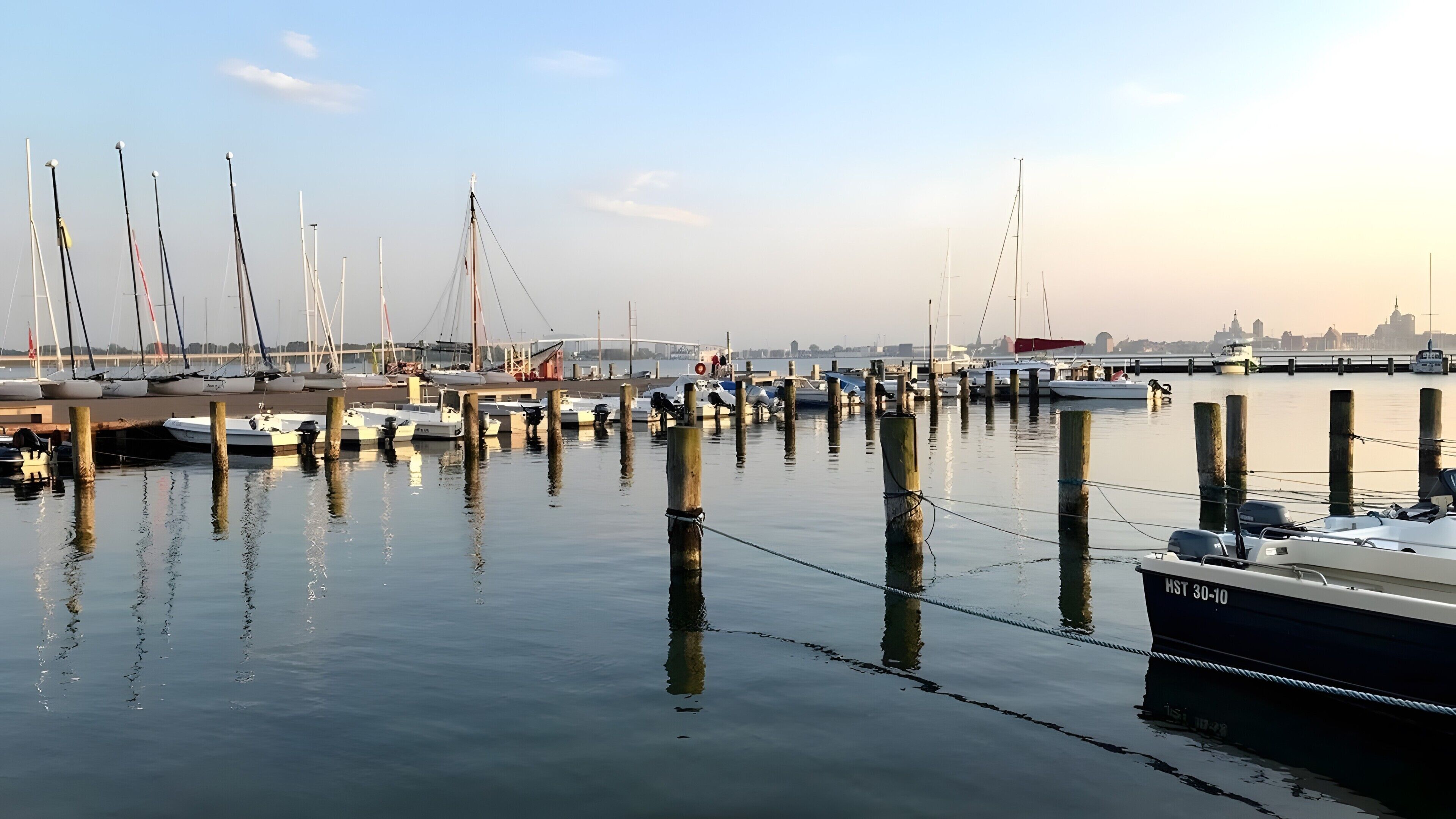Un port de plaisance où des bateaux sont amarrés à des poteaux en bois, l’eau est calme et, au loin, on aperçoit la silhouette de la ville.