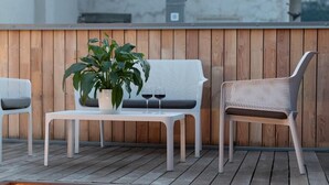Eine Holzterrasse mit einem weißen Tisch, zwei Korbstühlen, einem Blumentopf und zwei Weingläsern.