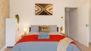 Une chambre à coucher comprenant un lit, une table de chevet, une lampe, un tableau ou une décoration murale, et une porte.