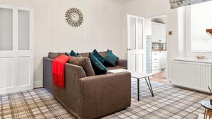 Un salon comprenant un canapé, une table basse, un tapis à motifs, une horloge murale et une fenêtre avec des stores.
