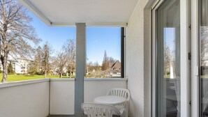 Varanda com mesa e cadeiras brancas, porta de vidro e vista para árvores e edifícios.