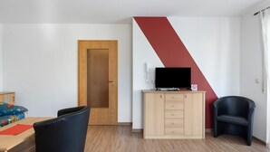 Quarto com porta de madeira, televisão sobre um móvel, uma cadeira preta e uma cama com roupa de cama colorida.