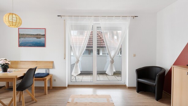 Et værelse med et træspisebord, en sort stol, en træbænk, en sort lænestol, et træskab, et billede på væggen og en skydedør med hvide gardiner.