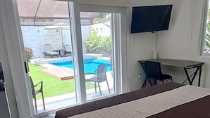 A hotel room with a bed, a desk with a chair, a TV, and a sliding glass door leading to a pool area. - Cozy private kingbed bungalow in peaceful Cancún with AC, Waterfall, Pool & Jets (Cancún)
