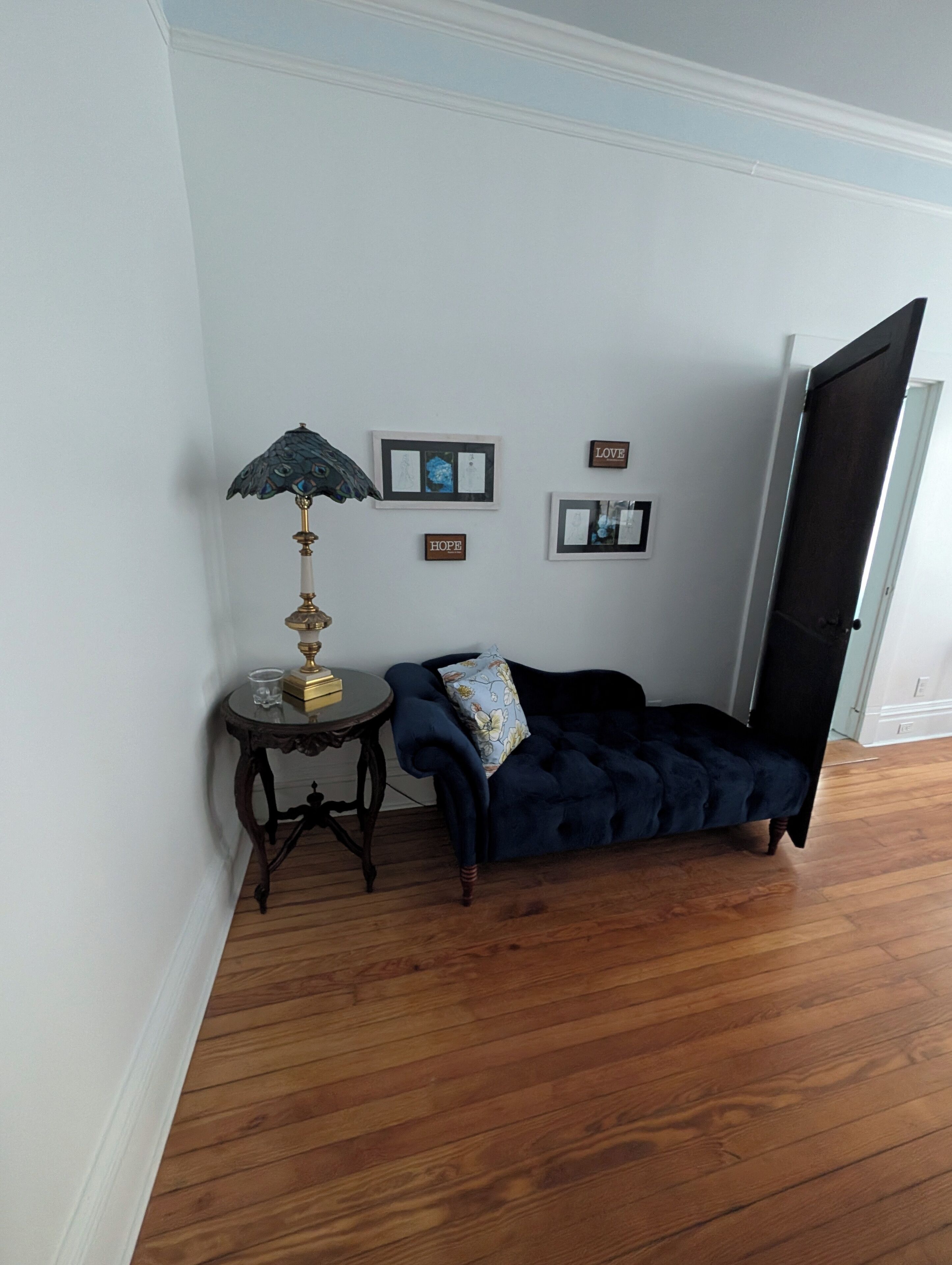 Una habitación con un sofá azul oscuro, una mesita redonda con una lámpara y cuadros en la pared.