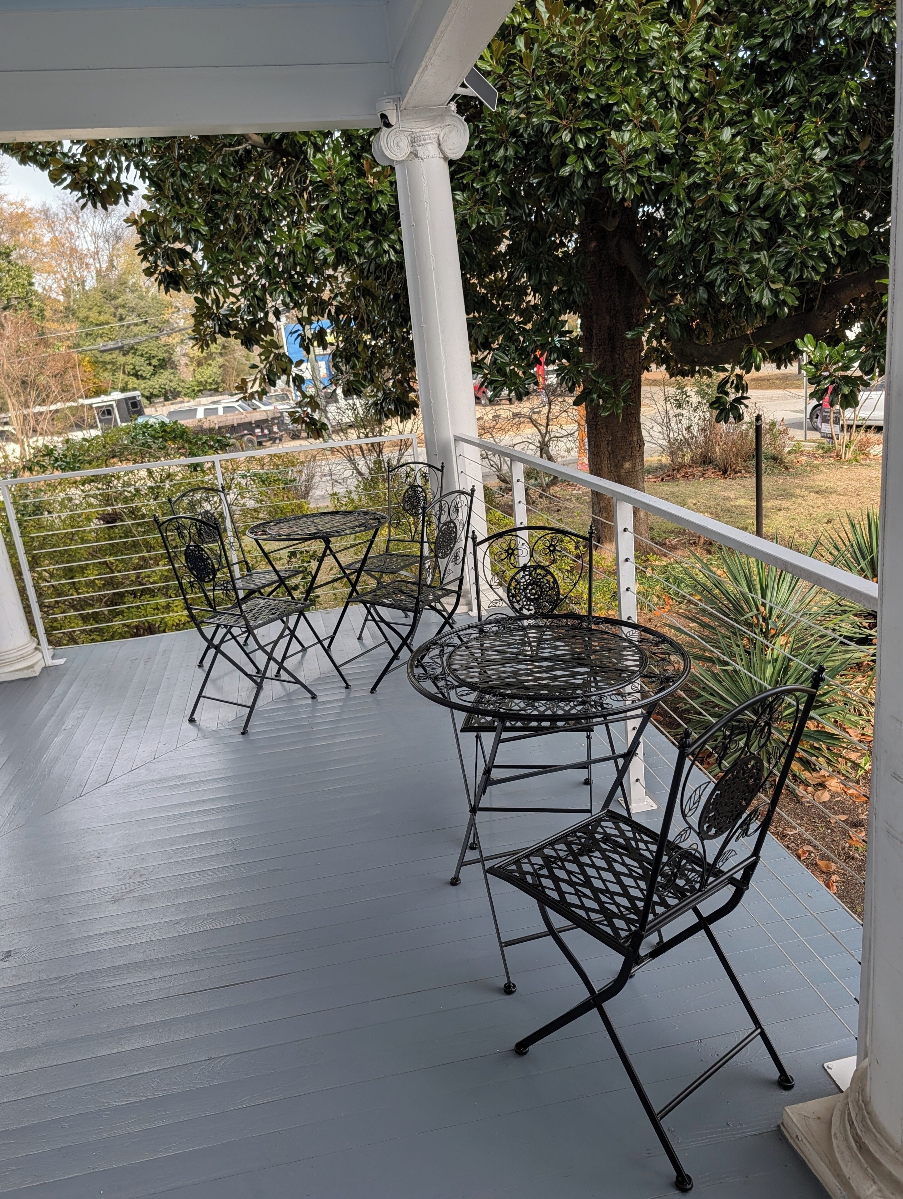 A covered outdoor seating area with metal tables and chairs, a white railing, and a tree in the background.