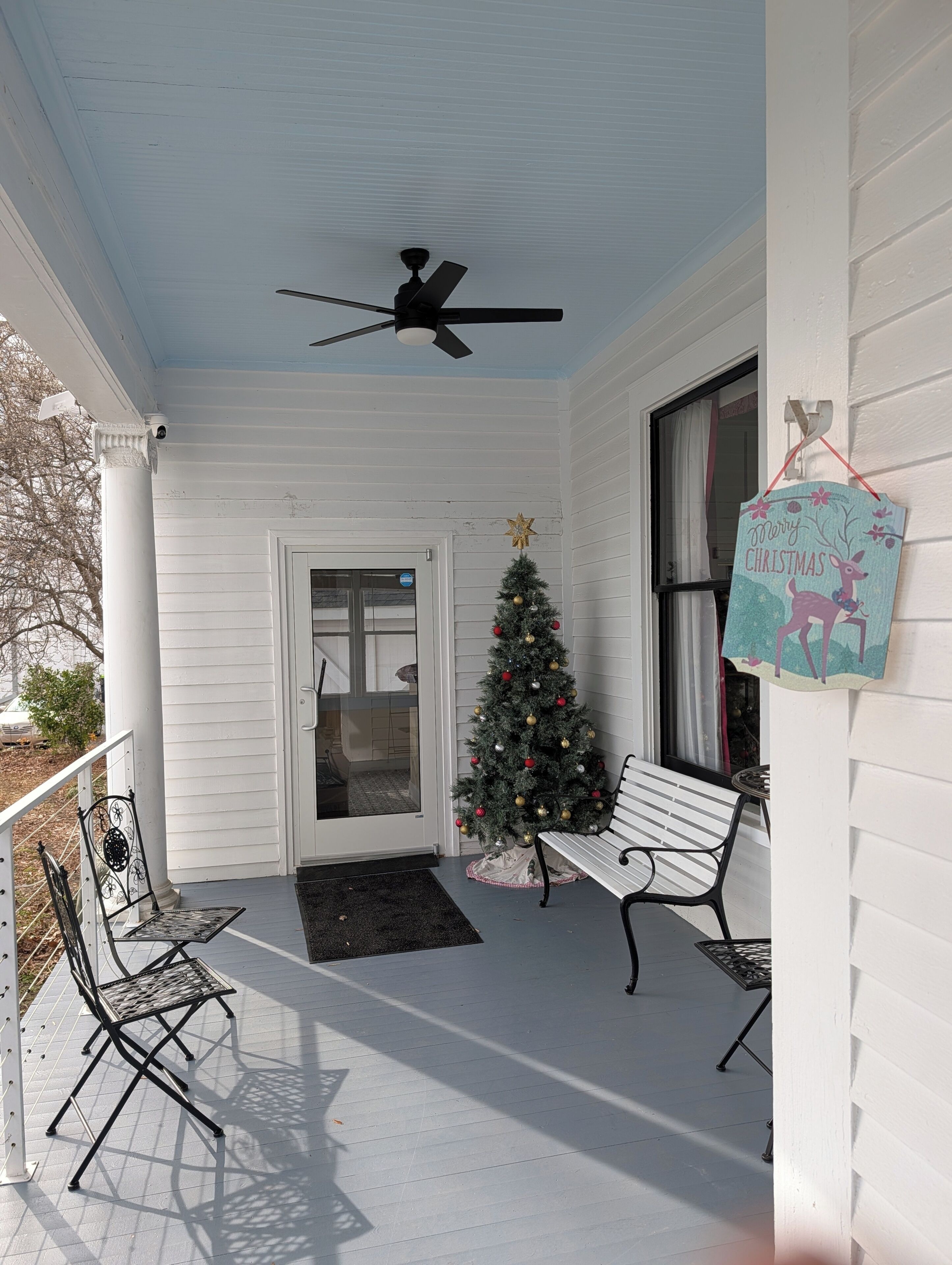 Un portico coperto con una ventola a soffitto, un albero di Natale, due sedie in metallo e una panchina.