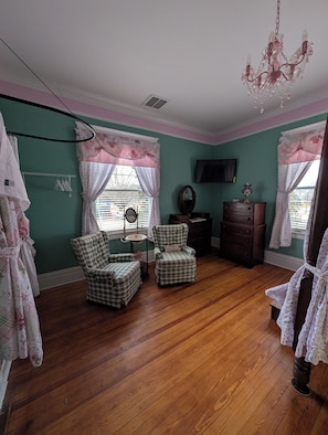 A room with teal walls, wooden flooring, two checkered armchairs, a dresser, a television, and a chandelier. - Grande Magnolia of Benson (Benson)