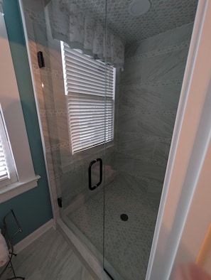 A glass-enclosed shower with a tiled floor and a window with blinds. - Grande Magnolia of Benson (Benson)