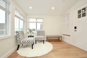 Ein Zimmer mit einer weißen Tür, einer Bank, zwei gemusterten Sesseln, einem weißen Teppich und einer Topfpflanze.